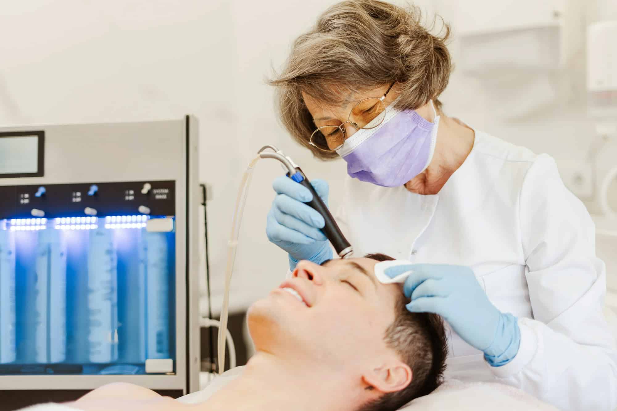 Senior woman cosmetologist performing mesotherapy on young, smiling man patient getting hydro facial